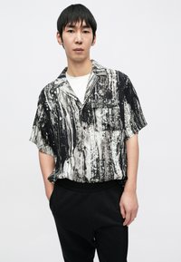 Kurzärmeliges Hemd in Schwarz und Weiß mit gestreiftem und gesprenkeltem Muster; leichter Stoff, Kragen und Knopfverschluss an einem Modell.