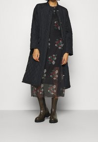 Svart vadderad långrock med hög krage, lager på lager över en mörk blommig klänning. Modell som bär mörkbruna gummistövlar med texturerad sula.