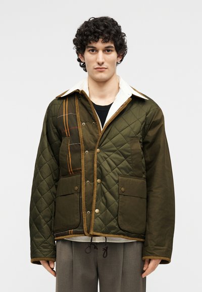 Νέος άνδρας με σγουρά μαλλιά φοράει απαλή τζάκετ ελιάς με τσέπες με επιδιορθώσεις και αντίθετο λευκό γιακά, στέκεται μπροστά σε απλό φόντο.