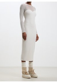 Robe blanche en tricot côtelé avec des manches longues et un col transparent, associée à des bottes beiges à lacets et des chaussettes en tricot crème.