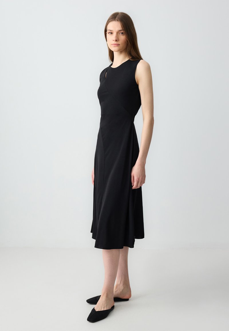 Ärmelloses schwarzes Kleid mit tailliertem Oberteil und ausgestellt Rock, aus weichem Stoff gefertigt. Verfügt über ein asymmetrisches Design und eine glatte Textur.