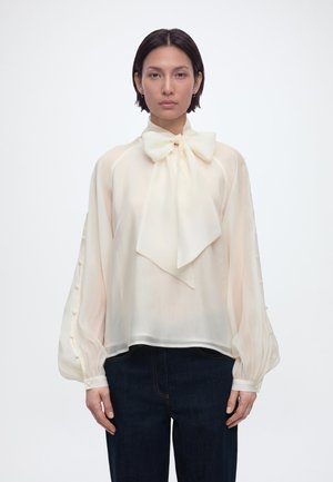 Woman wearing a sheer cream blouse with a large bow necktie, buttoned balloon sleeves, and dark pants, facing forward against a plain background.