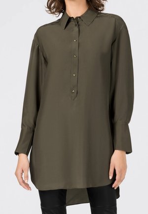 Femme portant une longue chemise boutonnée vert olive avec un col pointu et des manches longues, associée à un pantalon noir.