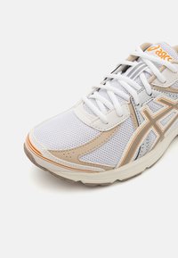 White athletic shoe with mesh upper, beige accents, orange detail, and gray stripes. Features laces, padded tongue, and textured sole.