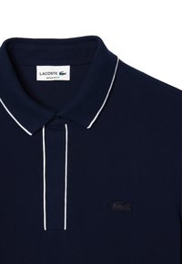 Marinblå pikétröja med texturerat tyg, vita accentstreck längs kragen och en liten svart krokodil-logotyp på bröstet.