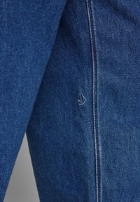 Tela de denim azul oscuro con una textura suave. Presenta un pequeño logotipo bordado y costuras visibles a lo largo de la costura.
