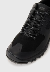 Black athletic shoe featuring a mix of smooth and textured materials, round laces with a diamond pattern, and a rugged outsole.