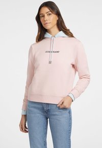 Rožinė megztinis su šviesiai mėlyna kapišonu, raukšlėtais rankogaliais ir apačia. Yra juodu išsiuvinėtas "GUESS JEANS" logotipas. Minkšta tekstūra.