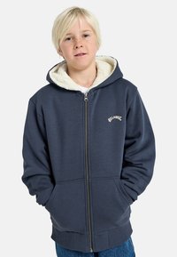 Felpa con zip blu navy, foderata in pelliccia sintetica bianca, con tasche frontali, logo ricamato crema, e polsini e orlo a costine.