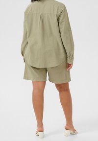 Licht olijfgroen, lichtgewicht shirt met drop shoulders, achterplooien en opgerolde mouwen, gecombineerd met bijpassende shorts. Gemaakt met witte hakken.