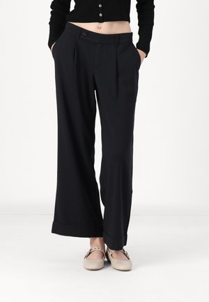 Kvinde iført sorte vidde-ben bukser, sort kort sweater og beige Mary Jane flade sko, stående med hænderne i lommerne på hvid baggrund.