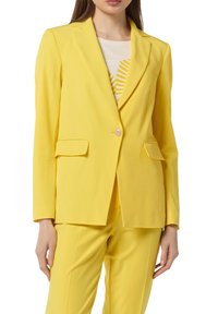 Blazer giallo con revers, chiusura a un bottone e due tasche frontali. Tessuto liscio; abbinato a un top bianco e giallo a fantasia.