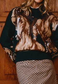 Γυναίκα που φοράει σκούρο πουλόβερ με μεγάλο καφέ και μπεζ ζωικό μοτίβο, συνδυασμένο με καφέ και λευκή καρό φούστα, στέκεται μπροστά σε ξύλινη πόρτα.