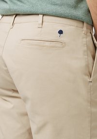 Pantalones de algodón beige con un bolsillo trasero plano. Presenta un pequeño logo de árbol en color marino bordado encima del bolsillo. Textura suave, ajuste entallado.