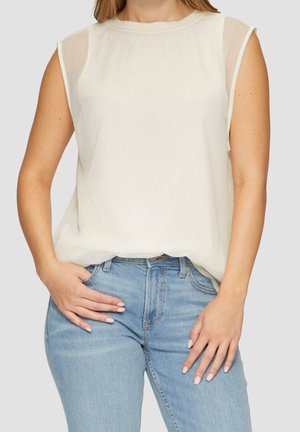 Woman wearing sleeveless white blouse with sheer shoulder panels and light blue high-waisted jeans, hands resting on hips.