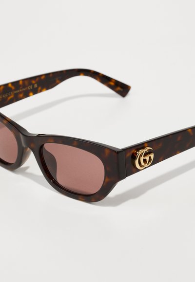 Lunettes de soleil écaille de tortue avec des verres teintés marron, présentant un accent doré du logo GG sur le côté. Design de cadre angulaire avec des branches larges.