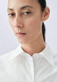 Weißes Hemd mit Button-Down-Front, glatte Textur, enganliegendes Design, getragen von einer Person mit minimalem Make-up und dunklem Haar.