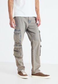 Pantaloni cargo in denim grigio chiaro, caratterizzati da molteplici tasche con zip e a patta. Indossati con sneakers marroni e una t-shirt bianca semplice.