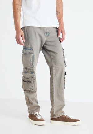 Cargo broeken in lichtgrijs denim, met meerdere rits- en klep pockets. G gedragen met bruine sneakers en een eenvoudig wit t-shirt.