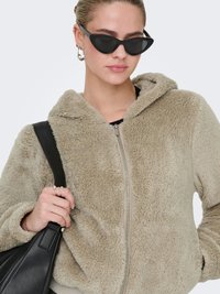 Beige fleece zip-up hoodie with a soft texture, featuring a hood and front pockets. Model wears black cat-eye sunglasses and holds a black bag.
