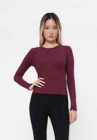 Donna con capelli lisci ombre che indossa un top a maniche lunghe in maglia aderente color porpora e pantaloni neri, in piedi davanti a uno sfondo bianco plain.