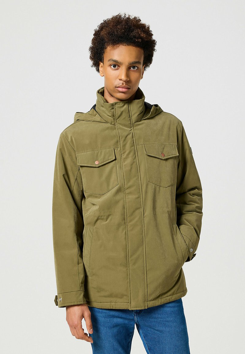 Oliwkowa kurtka zapinana na zamek z dwoma kieszeniami na piersi, odpinanym kapturem i gładką teksturą. Niebieskie dżinsy dopełniają całość.