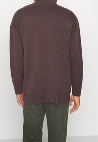 Dunkelbrauner gestrickter Pullover mit strukturiertem Rippmuster, Rundhalsausschnitt und langen Ärmeln. Abgeschlossen mit einem gerippten Saum und Bündchen. Trägt er über dunkelgrünen Hosen.