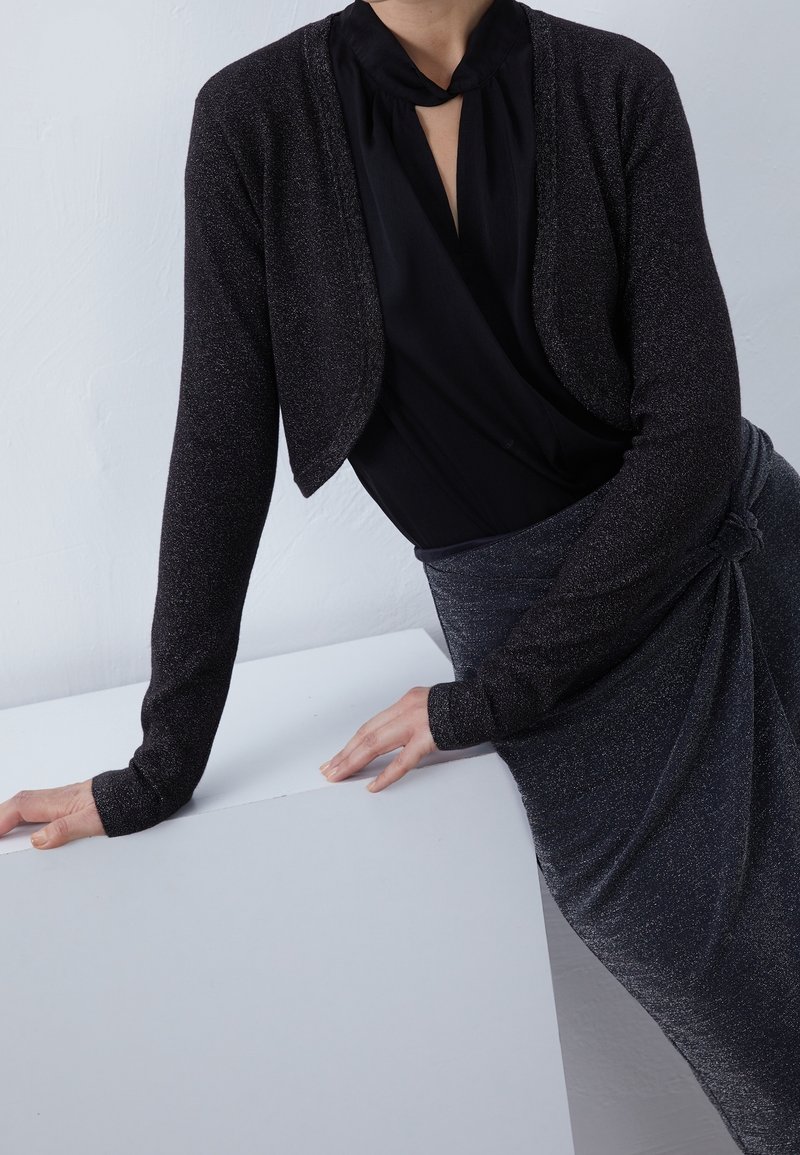 Femme portant un shrug noir scintillant à manches longues par-dessus un chemisier noir et une jupe scintillante assortie, s'appuyant avec les mains sur une surface blanche.