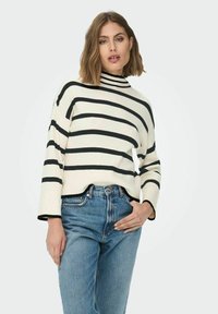 Balta un melna svītraina adīta džemperis ar augstu apkakli, brīvu piegriezumu un ribotu faktūru, kopā ar gaiši zilām džinsa biksēm.