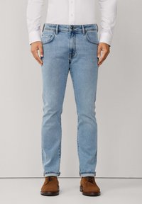 Muškarac u svijetloplavim uskim trapericama, bijeloj košulji na dugmad i smeškim suknjačama od suede, stoji uz jednostavnu pozadinu.