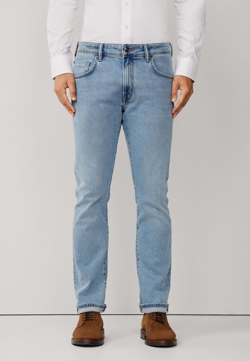 Muškarac u svijetloplavim uskim trapericama, bijeloj košulji na dugmad i smeškim suknjačama od suede, stoji uz jednostavnu pozadinu.