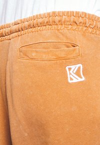 Orange sweatpants featuring a softly textured fabric, elastic waistband, and a small white logo patch near the back pocket.