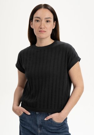 Woman with long dark hair wearing a black short-sleeve knit top and blue jeans, hands in pockets, against a plain light background.