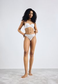 Soutien-gorge blanc rembourré avec des fines bretelles associé à un string avec bord en dentelle. Le mannequin est pieds nus sur un doux tapis gris contre un fond clair.