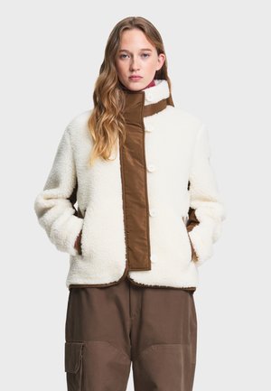 Giovane donna con capelli lunghi che indossa una giacca in pile bianca con dettagli marroni e pantaloni cargo marroni, in piedi con le mani nelle tasche.