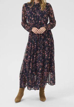 Kobieta w granatowej sukience w kwiatowy wzór z długimi rękawami, zapinanej na guziki z wiązaniem w talii, w połączeniu z beżowymi zamszowymi botkami do kostki, stojąca.