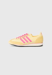 Gul atletisk sko med rosa detaljer. Har tre rosa striper, runde snøringer og en teksturert yttersåle. Mykt semsket skinn og syntetiske materialer.