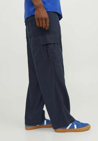 Pantalons cargo navy en tissu léger avec poches latérales. Associés à des chaussures de sport bleues avec des rayures blanches et une semelle en caoutchouc.