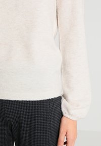 Lys beige sweater med blød tekstur og lange ærmer, parret med sorte ternede bukser med et subtilt gittermønster.