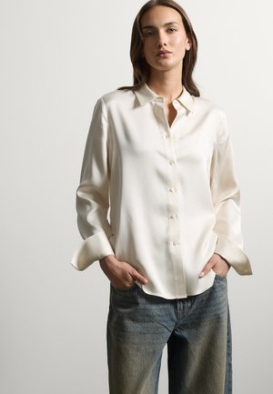 Donna con capelli castani che indossa una camicia bianca larga a maniche lunghe e jeans blu, in piedi con le mani in tasca.