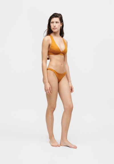 Femme debout pieds nus portant un bikini deux-pièces orange, légèrement tournée vers la droite sur un fond blanc uni.