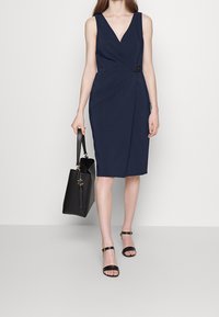 Marinblå ärmlös klänning med V-ringning, figursydd silhuett och en sidobälte detalj. Matchas med svart handväska och klacksandaler.