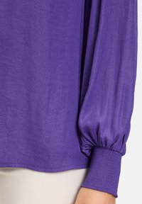 Lilla langærmet bluse med voluminøse, samlede ærmer og et tekstureret stof. Elastiske manchetter giver en tætsiddende afslutning ved håndleddene.