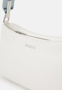Bílá kožená kabelka přes rameno se stříbrným zipem a světle modrým popruhem, s logem "HUGO" na přední straně v barvě stříbra.