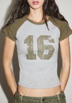 Junge Frau trägt ein graues und olivgrünes Baseball-T-Shirt mit einer großen abgenutzten Nummer 16 auf der Vorderseite.