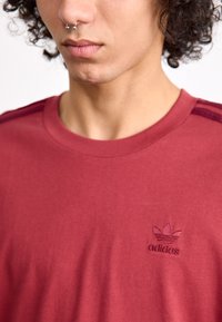Bordo spalvos crew neck marškinėliai su išsiuvinėtu Adidas logotipu ant krūtinės, su kontrastinėmis pečių juostelėmis panašiu atspalviu ir lygiu paviršiumi.