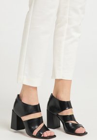 Schwarze Leder-High-Heel-Sandalen mit offenem Zeh, ausgestattet mit breiten Riemen und einem klobigen, quadratischen Absatz. Glatte Textur und minimalistisches Design.