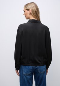 Sort silk bluse med krave, samlede manchetter og elastisk kant, parret med blå denim jeans. Glat tekstur og afslappet pasform.