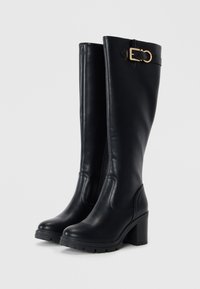 Bottes noires en cuir jusqu'au genou avec un talon épais et une boucle dorée décorative sur le côté. Présentent une texture lisse et une fermeture éclair latérale.