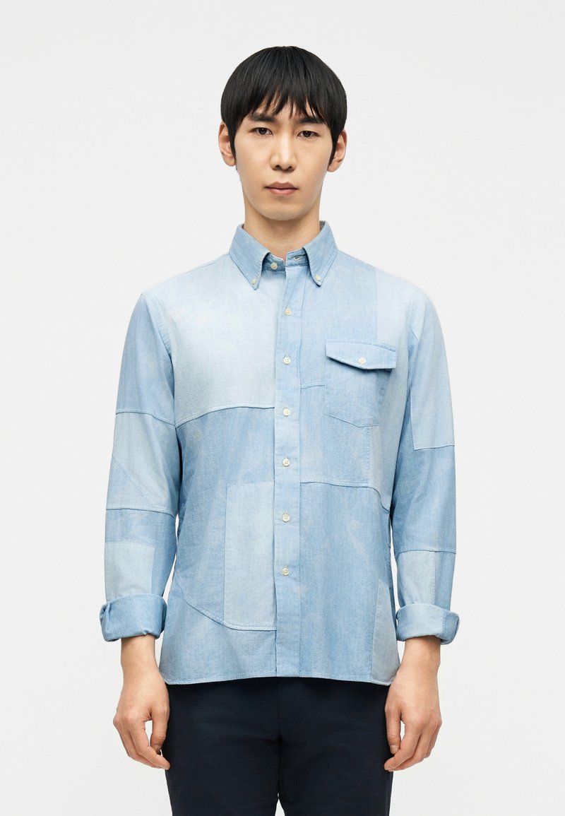 Άνδρας που φοράει μακρυμάνικο, κουμπωτό, κολάζ α νοιχτό μπλε τζιν πουκάμισο με μία κουμπωτή τσέπη στο στήθος και με γυρισμένα μανίκια.